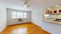 Located at 5651 N Magnolia Ave in Chicago's Edgewater neighborhood, this single-floor unit offers a clean, functional layout with consistent design elements throughout. Warm-toned hardwood flooring extends across the main living area and bedroom, lending continuity and a classic Chicago character to the space.

The open living area is bright and simply appointed, featuring white walls, a ceiling fan with light fixture, a baseboard heating system, and a wall-mounted air conditioning unit. Natural light enters through a window fitted with blinds, providing views of the surrounding neighborhood.

The kitchen is positioned adjacent to the living area and accessible via a pass-through counter, creating an open, connected feel. It is equipped with a gas range, refrigerator, under-cabinet range hood, white tile backsplash, speckled laminate countertops, and upper cabinets with wood-accented glass panels. A recessed light above the pass-through adds task lighting.

The bedroom is a comfortable, well-proportioned room with the same hardwood flooring, a ceiling fan, baseboard heat, and a window for natural light.

The bathroom features white mosaic tile flooring, a tub-shower combination with curtain, a vanity with sink, and a louvered door that opens to a built-in shelving unit, offering practical storage.