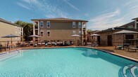 Located at 106 Pullman Lane in Mooresville, North Carolina, this single-story amenity building serves as a central hub for recreation and wellness. The exterior is characterized by neutral-toned siding, multi-pane windows, and a dark shingle roof, complementing the surrounding multi-story residential buildings.

The fitness center is a standout feature, accessible through sleek glass-paneled double doors. Large windows allow natural light to pour in, illuminating a well-appointed space equipped with strength training machines, free weights, and ceiling fans that maintain a comfortable environment during workouts. Adjacent to the fitness center is a dedicated yoga room, accessible via a glass door and marked with clear signage. A tiled accent wall and dual water fountains are conveniently positioned nearby.

The interior common areas feature polished concrete flooring and clean white walls, creating a contemporary and functional aesthetic. Large glass entry doors provide views of the surrounding residential courtyard.

Outdoors, the resort-style pool area is a highlight, offering a generously sized swimming pool with crystal-clear water. The surrounding deck is furnished with lounge chairs and shaded umbrellas, while a pergola-covered outdoor kitchen with grills and bar-height seating provides an ideal setting for social gatherings under open skies.