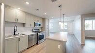 Located at 24000 W Lugonia Ave in Redlands, CA, this well-appointed single-floor residence offers a thoughtfully designed layout that maximizes comfort and functionality. The open-concept living area seamlessly connects the kitchen, dining, and living spaces, creating an inviting atmosphere filled with natural light from multiple windows. The modern kitchen is a standout feature, showcasing soft gray shaker-style cabinetry with matte black hardware, a generous island with a quartz countertop, stainless steel appliances including a gas range and built-in microwave, and stylish pendant lighting overhead. Light wood-tone flooring flows throughout the space, enhancing the bright and contemporary aesthetic. The spacious bedroom benefits from dual windows that invite natural light and features a ceiling fan fixture, two closets for ample storage, and direct access to the bathroom. The clean, minimalist bathroom includes a soaking tub with a curved black shower rod, matching gray cabinetry, and recessed lighting. Additional conveniences include an in-unit laundry area and a private balcony. Shaker-panel interior doors with matte black hardware complete the cohesive modern design found throughout this stylish Redlands residence.