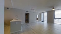 This contemporary single-floor space showcases a sleek, open-concept layout designed with modern living in mind. Light-toned hardwood flooring runs throughout, creating a seamless flow between the kitchen and living areas. The kitchen features a minimalist island with an integrated sink and a professional-style faucet, complemented by clean white surfaces that contribute to the space's bright, airy aesthetic.

Track lighting mounted on the ceiling provides adjustable illumination, while the open layout allows natural light to travel freely across the space. A floor-to-ceiling window and sliding glass door along one wall invite an abundance of daylight and offer direct access to an outdoor area, framed by vertical blinds for privacy and light control.

Neutral gray walls and white trim provide a refined, understated backdrop throughout. A hallway visible from the main living area leads to additional rooms, suggesting a well-organized floor plan. Ceiling-mounted HVAC vents are thoughtfully integrated into the design, maintaining the clean, uncluttered look. The overall ambiance is calm, contemporary, and move-in ready, with a minimalist design language that appeals to a wide range of lifestyles.