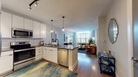 Located at 1300 N Fordham Blvd in Chapel Hill, NC, this well-appointed single-floor residence offers a thoughtfully designed open-concept layout with modern finishes throughout. The kitchen is a standout feature, showcasing crisp white cabinetry, a subway tile backsplash, quartz countertops, stainless steel GE appliances — including a refrigerator, range, microwave, and dishwasher — and a sleek black matte faucet at the central island, which also provides casual seating. Pendant lighting and track lighting add warmth and functionality to the space. The kitchen flows seamlessly into the living area, which is bathed in natural light from large windows with wood blinds. Wood-look flooring runs throughout the home, adding continuity and warmth. The primary bedroom features natural light from windows with sheer curtains, a walk-in closet with built-in shelving, and an en-suite bathroom with a glass-enclosed shower and a vanity with ample storage. A second bedroom also includes a walk-in closet and is served by a full hallway bathroom with a tub-shower combination and a single-sink vanity. The overall ambiance is contemporary and inviting, with a neutral color palette that provides a versatile backdrop throughout.