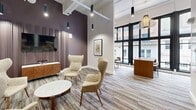Located in the heart of Hartford, Connecticut, this thoughtfully designed commercial space on Pratt Street offers a refined blend of contemporary style and functional versatility. The single-floor layout is anchored by soaring ceilings with exposed ductwork, lending an industrial-chic character that is softened by warm, curated interiors throughout.

The main lounge area features mid-century modern wingback chairs arranged around a marble-topped coffee table, set against a striking deep plum accent wall with vertical wood slat paneling and a mounted flat-screen television. Multicolored striped carpeting adds texture and visual warmth to the open floor.

Adjacent to the lounge, two dining areas offer distinct atmospheres. One is positioned beside dramatic floor-to-ceiling windows that flood the space with natural light and frame views of the surrounding urban streetscape. A built-in banquette with accent pillows and a round dining table create an intimate gathering spot beneath a sculptural woven pendant light. A second enclosed dining pod, framed by sage-green glass doors, provides a more private setting with its own warm-toned interior.

Pendant lighting throughout varies in style — from sleek cylindrical fixtures to decorative woven pendants — creating layered illumination across the space. A green-framed glass entry door with direct street access completes this welcoming and well-appointed commercial environment.