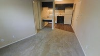 This single-floor space features an efficient, open-concept layout that connects the living area directly to the kitchen, maximizing usability within a compact footprint. The living area is finished with neutral-toned carpet, providing a comfortable and understated base for furnishings. A smooth transition leads into the kitchen zone, where the flooring shifts to warm hardwood-style planks that add a touch of contrast and durability underfoot. The kitchen is equipped with dark espresso cabinetry that extends from base to upper units, offering ample storage. Essential appliances are in place, including a white refrigerator, a stove, and a sink, all neatly arranged along the kitchen perimeter. A peninsula-style counter with a dark surface extends into the space, serving as a natural divider between the living and kitchen areas while also providing additional prep or dining space. A doorway visible from the main living area suggests access to additional rooms or storage. The overall ambiance is clean and functional, with neutral wall tones and white trim throughout that keep the space feeling bright and move-in ready.