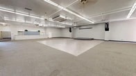 Located at 4300 Pan American Fwy in Albuquerque, New Mexico, this single-floor commercial space offers a generous and highly flexible layout well-suited for a variety of uses, from events and gatherings to office or training functions. The space is organized into two primary open areas that flow together to create an expansive, unobstructed floor plan.

The main hall is a standout feature, boasting a combination of light wood-style vinyl flooring and carpeted areas that help define distinct zones within the open layout. A low-profile raised stage provides a natural focal point for presentations or performances. Overhead, rows of fluorescent linear lighting ensure bright, even illumination throughout, complemented by ceiling fans and a ceiling-mounted projector for audio-visual presentations. HVAC units are integrated into the ceiling, ensuring climate control across the entire space.

A built-in reception or service counter with overhead pendant lighting anchors one section of the space, offering practical functionality for check-in or administrative use. Wide, carpeted corridors connect the main areas, with tall vertical windows drawing in natural light. White walls and dark baseboards create a clean, neutral aesthetic throughout. The textured ceiling adds a subtle architectural detail, completing this well-equipped and adaptable commercial environment.