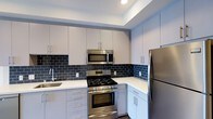 This single-floor residence showcases a thoughtfully designed interior with a strong emphasis on modern aesthetics and functional living. The kitchen is a standout feature, boasting a clean, contemporary layout with flat-front cabinetry in a soft grey-white finish, complemented by brushed silver hardware. A bold dark navy subway tile backsplash adds a striking contrast to the bright white countertops, creating a visually dynamic focal point. The kitchen is fully equipped with a full suite of stainless steel appliances, including a gas range with oven, an over-the-range microwave, and a full-size refrigerator, making it as practical as it is attractive. Recessed lighting illuminates the space efficiently, enhancing the clean lines and polished surfaces throughout. The cabinetry extends to the ceiling, maximizing storage while reinforcing the sleek, uncluttered look. The space reflects a cohesive modern design sensibility, with attention to detail evident in every finish and fixture. The overall ambiance is bright, functional, and sophisticated, offering a comfortable and stylish environment suited to contemporary living.
