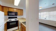 This single-floor unit is designed with a practical, straightforward layout that maximizes functionality across its living spaces. The kitchen is efficiently arranged in an L-shaped configuration, featuring warm honey-toned oak cabinetry that extends to the upper and lower walls, providing generous storage throughout. Appliances include an electric range, built-in microwave, dishwasher, and refrigerator, all complemented by light-toned countertops and tile flooring. A structural column partially separates the kitchen from the main living area, creating a subtle visual distinction between the two spaces.

The living area is finished with neutral gray carpet and benefits from a large window dressed with vertical blinds, allowing natural light to fill the room while offering an outdoor view. The space flows openly toward a hallway that connects to the remaining rooms, including a closet with ample storage.

The bathroom is clean and functional, featuring a full tub-shower combination with a sliding enclosure, a single vanity with an oak cabinet base and mirror above, and light vinyl flooring. The overall color palette throughout the unit is neutral, with white walls and consistent warm wood tones, lending the space a cohesive and understated aesthetic well-suited for comfortable everyday living.