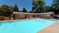 This community pool area offers a well-appointed outdoor aquatic facility designed for both recreation and lap swimming. The generously sized swimming pool features clearly marked lane dividers, making it suitable for leisurely swims and structured exercise alike. The pool's vivid blue water is complemented by a broad concrete deck that surrounds the perimeter, providing ample space for lounging and socializing. Blue and white lounge chairs and Adirondack-style seating are positioned throughout the deck, offering comfortable spots to relax under the open sky. A low-profile clubhouse-style building runs along one side of the pool, housing separate men's and women's restroom facilities accessed via dedicated exterior doors, adding a layer of convenience for pool users. A storage structure with vertical panel siding is also situated adjacent to the building. The pool area is enclosed by a metal security fence, ensuring a safe and contained environment. Mature deciduous and evergreen trees frame the space on all sides, providing natural shade, privacy, and a lush green backdrop against the clear blue sky. Lamp posts are positioned around the deck, suggesting the area is usable during evening hours as well.