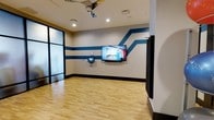 This yoga room is a purpose-built wellness space designed to facilitate guided movement and fitness sessions. The room features smooth hardwood flooring that provides a clean, resilient surface ideal for yoga, stretching, and exercise routines. The walls are finished in a neutral tone and accented with bold teal horizontal and diagonal stripes, giving the space a dynamic, energetic character that reinforces its active-use purpose. A large wall-mounted flat-screen display is positioned at the front of the room, enabling participants to follow along with instructional video content. A smaller wall-mounted touchscreen panel nearby offers convenient control over the room's media and settings. Recessed ceiling lighting provides bright, even illumination throughout the space, while a ceiling fan supports air circulation during active sessions. A glass-panel partition wall along one side allows for natural visibility into or out of the space, contributing a sense of openness. A wall-mounted accessory holder on the opposite side adds a functional storage element. Overall, the room is thoughtfully outfitted to deliver a comfortable, tech-enabled environment for individual or group wellness activities.