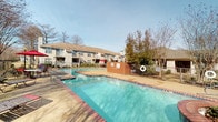 This outdoor pool area offers a well-appointed retreat designed for relaxation and leisure. The centerpiece is a generously sized swimming pool with clear, blue water framed by brick coping and accessible via handrail-equipped entry steps. The surrounding sundeck is finished in a textured aggregate concrete surface, providing ample space for lounging and socializing. Metal lounge chairs are positioned along the perimeter, offering spots to enjoy the open sky and natural light throughout the day. A dedicated patio seating area features dining tables paired with red accent chairs and shaded by bold red market umbrellas, creating a comfortable space for outdoor gatherings. Decorative lampposts are placed along the deck, providing illumination for evening use. The area is enclosed by a wrought iron fence with a safety life ring mounted at the gate, ensuring security while maintaining visibility to the surrounding landscape. Mature trees and greenery border the exterior of the fenced perimeter, offering a natural backdrop and a sense of seclusion. Landscaped planting beds with low shrubs add a polished, maintained appearance to the space. The multi-story residential buildings visible in the background complete the community setting of this outdoor amenity.