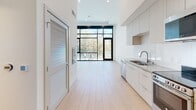 This contemporary single-floor space showcases a clean, minimalist design characterized by crisp white walls, light hardwood flooring, and soaring ceilings that lend an airy, expansive feel throughout. The open-concept layout seamlessly connects the kitchen and living area, creating a cohesive and functional flow. The kitchen is well-appointed with flat-front white cabinetry extending to the ceiling, stainless steel appliances including a dishwasher, electric range, microwave, and refrigerator, along with a double stainless steel sink set into a sleek white countertop. A striking floor-to-ceiling black-framed window wall anchors the living area, flooding the space with abundant natural light and offering direct views of lush greenery beyond a private outdoor area. The main room features impressively tall ceilings and includes a built-in closet with shelving, offering practical storage without compromising the unit's open character. The bathroom is finished in neutral tones with light flooring, accessibility grab bars, and clean, understated fixtures. Throughout the space, recessed lighting, modern ventilation, and consistent design details reinforce a polished, contemporary aesthetic suited for a variety of uses.