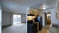 This single-floor space offers a practical and functional layout centered around an open-concept design that connects the kitchen and living area seamlessly. The kitchen is equipped with black appliances, including a coil-burner range and a full-size refrigerator, complemented by light wood cabinetry above and a range hood for ventilation. A granite-style countertop adds a touch of character to the cooking space, while tile flooring grounds the kitchen zone. The living area transitions into plush carpeting, creating a comfortable and cozy atmosphere for relaxing or entertaining. A sliding glass door floods the living space with natural light and provides direct access to a private balcony, offering views of the surrounding exterior. The textured ceiling runs throughout the space, adding a classic residential touch. A hallway visible from the main living area suggests additional rooms beyond, contributing to the overall functional flow of the layout. The open sightlines between the kitchen and living room create an inviting, connected feel throughout the home.