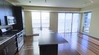 This single-floor space showcases a sleek, contemporary design centered around an open-concept layout that seamlessly connects the kitchen and living areas. The kitchen is anchored by a substantial dark-finished island with a smooth countertop surface, complemented by stainless steel appliances and dark cabinetry, creating a striking contrast against the warm-toned walls. Rich hardwood flooring runs throughout, adding warmth and continuity to the space.

Natural light floods the interior through multiple large windows and a floor-to-ceiling sliding glass door fitted with vertical blinds, framing expansive urban views that suggest an elevated position within the building. The sliding door leads to an outdoor area, extending the living space beyond the interior. The open living zone beyond the kitchen offers a versatile, unobstructed area well-suited for a variety of furniture arrangements. Recessed and track lighting supplements the abundant natural light. The overall ambiance is clean, modern, and urban, with a neutral color palette that provides a refined yet functional living environment.