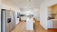 Located at 9000 Little Palm Way in Venice, FL, this well-appointed single-story home offers 2 bedrooms, 2 bathrooms, and a thoughtfully designed open-concept layout. The kitchen is a standout feature, showcasing crisp white shaker cabinetry, a grey subway tile backsplash, stainless steel appliances, quartz countertops, and a central island with seating — ideal for casual dining and entertaining. It flows seamlessly into the bright living room, which is bathed in natural light and finished with light wood-tone flooring that runs throughout the home. Both bedrooms are comfortably sized, with the primary bedroom offering ample space, and each room features windows that invite natural light. The two bathrooms are finished with white cabinetry, quartz vanities, and tiled tub-shower combinations. A dedicated laundry room with a full-size washer and dryer adds everyday convenience. Three closets provide generous storage throughout. A hallway connects the private quarters with clean, neutral-toned walls enhancing the airy feel. Outside, a private fenced yard with lush artificial turf and a patio area offers a low-maintenance outdoor retreat. The home's light blue lap siding exterior complements the serene Florida surroundings.