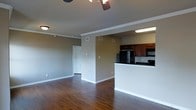 This single-floor residence showcases an open-concept layout that seamlessly connects the living area to the kitchen. The living space is finished with rich, dark hardwood floors that extend throughout, adding warmth and character to the interior. Crown molding lines the ceiling, providing a clean, polished detail, while a ceiling fan with a light fixture serves as a functional centerpiece overhead.

The neutral, warm-toned walls create a versatile backdrop well-suited to various décor styles. A doorway visible from the main living area leads to additional rooms beyond, hinting at a practical and connected floor plan.

The kitchen is defined by a raised breakfast bar that partially separates it from the living space while maintaining an open feel. Dark-stained upper cabinetry pairs with black appliances, including a refrigerator and range, lending a cohesive, contemporary look. Under-cabinet and overhead fluorescent lighting ensures the kitchen area remains well-lit. The overall ambiance of the space is clean and functional, with classic architectural details that give it a timeless residential character.