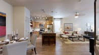 This single-floor residence offers a thoughtfully designed open-concept layout that seamlessly connects the kitchen, dining, and living areas into one cohesive living space. The kitchen is a standout feature, showcasing warm honey-toned cabinetry, stainless steel appliances including a refrigerator, microwave, and range, and a dark granite-topped central island with bar seating for two — ideal for casual dining or entertaining. A track lighting system and elegant pendant lights illuminate the kitchen, adding both function and visual interest. The dining area, positioned just off the kitchen, provides an intimate setting with upholstered seating and a coordinating light fixture. Wood-tone laminate or hardwood flooring flows continuously throughout the space, lending warmth and a cohesive aesthetic. The living area is bathed in natural light from large windows dressed with neutral drapes, creating a bright and welcoming atmosphere. A ceiling fan adds comfort to the space, which is styled with neutral-toned furnishings, layered textiles, and tasteful wall art. The overall ambiance is warm, modern, and inviting, with a design that balances everyday functionality with refined comfort.