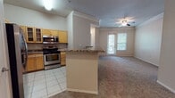 This single-floor residence offers a well-organized layout with a seamless flow between its key living spaces. The kitchen is equipped with stainless steel appliances, including a refrigerator, electric range, and over-the-range microwave, complemented by warm-toned wood cabinetry with glass-front accents, a tile backsplash, and granite-style countertops that extend across a peninsula breakfast bar. Tile flooring grounds the kitchen area, which opens directly into the carpeted living room. The living room is a comfortable, open space highlighted by crown molding, a ceiling fan, a built-in arched shelving nook, and a door providing access to the outdoors and natural light. Multiple bedrooms feature neutral carpet, large windows with blinds that allow ample natural light, and clean, minimalist walls offering versatile use of space. Two bathrooms are present — one featuring wood-look luxury vinyl flooring, a granite-top vanity, and a tub-shower combo with a distinctive oval window above, and a second with a granite countertop vanity and a tub-shower combination. A hallway connects the living areas to the private rooms, completing the functional and cohesive floor plan.