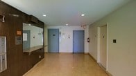 This single-floor space presents a practical and well-maintained common area, likely serving as a building lobby or transitional hub. The layout is open and uncluttered, with a spacious central floor area finished in speckled terrazzo tile that lends a classic, durable character to the space. Recessed ceiling lighting provides even, consistent illumination throughout, contributing to a bright and functional atmosphere. A built-in reception or service counter runs along one side of the space, constructed with a dark wood-toned finish and featuring a glass or mirrored partition above it, suggesting a security or administrative function. The far wall features two prominent blue-grey doors that serve as elevator entrances, each equipped with call panels and informational signage. Safety compliance is evident with a mounted fire extinguisher positioned between the elevator doors. A third door to the right leads to an adjacent corridor or stairwell, extending the accessibility of the space. The walls are finished in a neutral off-white tone, and the overall design prioritizes efficiency and ease of movement. The space conveys a straightforward, institutional character well-suited to a commercial or multi-unit residential building.