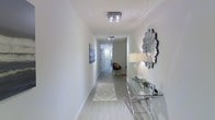 This single-floor space showcases a refined modern aesthetic defined by clean lines, neutral tones, and thoughtful design details. The entryway immediately establishes the character of the space — a long, airy corridor with crisp white walls, light hardwood-style flooring, and flush-mount crystal ceiling fixtures that cast a bright, even glow. A decorative sunburst mirror adorns one wall, accompanied by a sleek glass-and-chrome console table that adds a sophisticated, contemporary touch. A geometric-patterned area rug grounds the hallway, guiding visitors deeper into the space. The corridor connects to additional rooms at the far end, hinting at an open and functional layout beyond. Natural light filters through the space, enhancing the bright, clean ambiance created by the white walls and pale flooring. The overall design language is minimalist yet warm, balancing modern furnishings with subtle decorative accents. The high-quality finishes and attention to detail throughout suggest a well-appointed interior that is both stylish and livable, making this a polished and inviting space suited to a variety of uses.