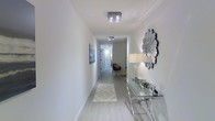 This single-floor space showcases a refined modern aesthetic defined by clean lines, neutral tones, and thoughtful design details. The entryway immediately establishes the character of the space — a long, airy corridor with crisp white walls, light hardwood-style flooring, and flush-mount crystal ceiling fixtures that cast a bright, even glow. A decorative sunburst mirror adorns one wall, accompanied by a sleek glass-and-chrome console table that adds a sophisticated, contemporary touch. A geometric-patterned area rug grounds the hallway, guiding visitors deeper into the space. The corridor connects to additional rooms at the far end, hinting at an open and functional layout beyond. Natural light filters through the space, enhancing the bright, clean ambiance created by the white walls and pale flooring. The overall design language is minimalist yet warm, balancing modern furnishings with subtle decorative accents. The high-quality finishes and attention to detail throughout suggest a well-appointed interior that is both stylish and livable, making this a polished and inviting space suited to a variety of uses.