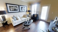 This well-appointed single-floor residence exudes a refined yet welcoming ambiance, thoughtfully designed with both comfort and style in mind. The living area serves as the heart of the space, showcasing rich, dark hardwood flooring that adds warmth and a sense of understated luxury throughout. Crown molding along the ceiling lends an architectural elegance that complements the neutral, warm-toned walls.

Natural light fills the room generously, pouring in through large windows dressed in sheer curtains and slatted blinds, as well as a full-length glass door that opens to the outside. This seamless connection between interior and exterior creates an airy, open feel. A crisp white sofa anchored by decorative throw pillows pairs gracefully with a sleek black accent chair, while an oval glass coffee table adds a contemporary touch to the seating arrangement.

The walls are adorned with framed black-and-white botanical artwork, contributing a sophisticated gallery-like quality to the living space. Overhead track lighting provides additional illumination, ensuring the space is equally inviting in the evening hours. Overall, this residence offers a harmonious blend of classic design elements and modern sensibility, making for a polished and comfortable living environment.