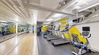 This fitness center offers a comprehensive and thoughtfully designed workout environment spread across a single floor. The space is defined by its bold aesthetic — striking geometric patterns in yellow, white, and gray cover the walls, lending the facility a modern and energetic atmosphere that motivates movement throughout.

The cardio zone is well-appointed, featuring a row of treadmills, elliptical trainers, and stationary bikes, all equipped with individual display screens. Adjacent to the cardio area, a dedicated strength training section includes a cable machine, a dumbbell rack with a weight bench, and additional resistance equipment — providing a well-rounded selection for full-body training.

A standout feature of the space is a dedicated studio area with warm hardwood flooring, full-length mirrored walls, and a wall-mounted ballet barre, making it ideal for dance, yoga, stretching, or group fitness sessions. This section creates a clean visual contrast against the rubber flooring of the main gym area.

The ceiling is open and industrial in character, with exposed ductwork and pipework painted white, contributing to the facility's contemporary feel. Overhead fluorescent lighting ensures the space is evenly and brightly lit throughout. Small windows along the upper walls allow for natural light to supplement the interior lighting, adding to the overall openness of the environment.