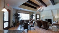 This inviting single-floor gathering space showcases a warm, Spanish Colonial-inspired aesthetic that combines rustic charm with refined comfort. The ceiling is a standout architectural feature, adorned with bold dark wood exposed beams set against a coffered tray design, complemented by an elegant wrought-iron chandelier that anchors the room with a classic, old-world ambiance. Terra cotta tile flooring runs throughout, reinforcing the space's warm, earthy character and lending a sense of cohesion across the open layout. Large windows dressed with neutral drapes allow an abundance of natural light to flood the interior, creating a bright and airy atmosphere while offering views of the surrounding greenery. The space is thoughtfully arranged with multiple seating areas, including a circular dining table with dark leather chairs, as well as wicker and upholstered lounge seating, accommodating both casual conversation and more structured gatherings. Lush indoor plants are strategically placed throughout, enhancing the organic, welcoming feel of the room. A fireplace and built-in shelving along one wall add further architectural interest and functionality. Overall, the space strikes a balance between communal functionality and residential warmth, making it well-suited as a lounge, clubroom, or common area.