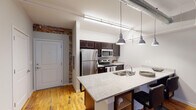 Located at 223 Virginia St in Mount Airy, North Carolina, this single-floor unit showcases a compelling industrial-chic aesthetic achieved through the thoughtful conversion of a historic building. Original exposed brick walls are a defining architectural feature found throughout the space, adding texture and character to nearly every room. Warm hardwood floors run continuously underfoot, complementing the raw industrial elements above, including visible ductwork and structural details at the ceiling.

The open-concept kitchen serves as a functional centerpiece, featuring a large granite-topped island with bar seating, dark espresso cabinetry, stainless steel appliances, and pendant lighting suspended from above. The living area benefits from generous ceiling height, exposed piping, and a tall multi-pane window set within a brick archway that fills the room with natural light.

Multiple bedrooms are present, each with its own character. One features full brick walls on multiple sides with a bright window and bunk bed setup, while another offers a quieter, more refined atmosphere with softer gray walls and exposed ductwork overhead. The bathroom is clean and modern, with granite-topped vanity, dark cabinetry, and a full tub-shower combination, maintaining consistent finishes found throughout the space.