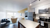 This single-floor unit showcases a thoughtfully designed open-concept layout that seamlessly connects the kitchen, dining, and living areas. The kitchen is equipped with stainless steel appliances, including a full-size refrigerator and dishwasher, complemented by white upper cabinetry and warm wood-toned lower cabinets. A spacious concrete-finish dining table anchors the central space, offering both function and style. Track lighting overhead provides focused illumination throughout.

The living area features warm hardwood-style flooring and neutral tones accented by a yellow feature wall, creating a welcoming atmosphere. A pair of modern swivel chairs and a dresser complete the living space, while large windows allow natural light to flow in. A separate bedroom is visible through an open doorway, furnished with a bed and offering a degree of privacy from the main living areas. The overall aesthetic is clean and contemporary, with a cohesive palette of whites, warm wood tones, and soft yellows that give the space a bright, comfortable feel.
