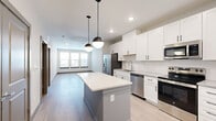 This well-designed one-bedroom, one-bathroom residence offers a thoughtful layout across a single floor, combining modern finishes with functional living spaces. The open-concept kitchen and living area serves as the heart of the home, featuring crisp white shaker-style cabinetry, a full suite of stainless steel appliances including a French door refrigerator with ice maker, dishwasher, range, and microwave. A center island with a white quartz countertop provides additional prep space and casual seating, accented by stylish pendant lighting overhead. Light wood-style luxury vinyl flooring flows seamlessly throughout the entire residence, lending a clean, cohesive aesthetic. The living area benefits from natural light through large windows, creating an open and airy atmosphere. The bedroom is a well-proportioned retreat equipped with a ceiling fan and a window with exterior views, offering comfortable natural light. An adjacent walk-in closet provides generous storage with built-in shelving and hanging rods. The full bathroom features a white vanity with quartz countertops, a large framed mirror, black hardware accents, and white subway tile detailing, connecting directly to the bedroom for added convenience. The overall design palette — neutral tones, clean lines, and contemporary fixtures — creates a polished, move-in-ready living environment.