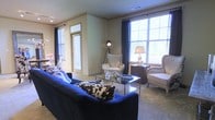 This single-floor residence offers a thoughtfully designed living space that balances comfort and sophistication. The living area serves as the centerpiece of the home, showcasing large, multi-pane windows dressed with floor-length navy drapery that frame generous views of the outdoors while allowing an abundance of natural light to fill the room. Crown molding adds a refined architectural detail along the ceiling line, complementing the neutral wall tones throughout.

The living area is furnished with a deep navy sofa accented by bold patterned throw pillows, paired with classic wingback chairs in a neutral upholstery. A glass-top coffee table anchors the seating arrangement, reflecting light and contributing to an open, airy feel. A traditional table lamp and a sculptural arc floor lamp provide layered ambient lighting. An entryway area with additional windows and a glass-paneled door is visible from the main living space, reinforcing the home's bright and welcoming atmosphere.

The overall ambiance of the space is elegant yet approachable, with a curated mix of classic furnishings, decorative accents, and architectural detailing that creates a cohesive and inviting interior environment suitable for both everyday living and entertaining.
