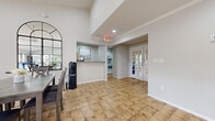 This single-floor community space is thoughtfully designed to accommodate a variety of functions within one cohesive layout. The interior is divided into clearly defined zones, each with its own character while maintaining a unified, contemporary aesthetic throughout.

The lounge area makes a strong visual impression with its dramatic shiplap accent wall in crisp white, complemented by light gray wood-look flooring. A fireplace console with barn-door hardware serves as the focal point beneath a wall-mounted television. Tall floor-to-ceiling windows flood the space with natural light and provide open views of lush, palm-lined outdoor surroundings. Comfortable seating and a workspace area complete this versatile zone.

The dining area features vaulted ceilings with a ceiling fan, warm tan tile flooring, and arched decorative mirrors adorning the walls. Multiple dining tables with upholstered chairs accommodate groups of varying sizes, while a water station adds practical convenience. The space benefits from natural light through multiple windows and a French door leading outdoors.

The kitchen is efficiently arranged with white cabinetry, stone-look countertops, white appliances including a range and refrigerator, and subway tile backsplash. A pass-through counter connects it to the main gathering areas. The overall ambiance throughout is bright, welcoming, and well-appointed.