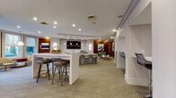 This thoughtfully designed single-floor amenity lounge offers a seamless blend of social, recreational, and work-oriented spaces within a contemporary interior. The space is anchored by a large central white island with bar seating, complemented by a secondary stone-accented bar area equipped with additional counter seating and a wall-mounted flat-screen TV — ideal for casual gatherings. Warm wood-paneled accent walls contrast elegantly with light-toned carpeting featuring a geometric diamond pattern and natural wood-look flooring near the entrance.

A dedicated co-working alcove provides a row of individual workstations, each framed by light wood partitions and outfitted with sleek black swivel chairs and pendant lighting, creating a focused yet stylish work environment. The lounge area near large multi-pane windows is bathed in natural light and offers views of an outdoor courtyard, furnished with a pair of cream swivel chairs and a metallic accent table beneath a statement pendant lamp.

Recreational highlights include a billiards table, a shuffleboard table, and a wall-mounted oversized Scrabble board set against a rich wood-paneled nook. The entrance features striking deep blue double doors flanked by warm wood accents and polished recessed lighting, creating a welcoming and refined first impression throughout.