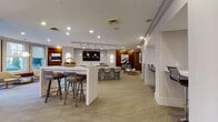 This thoughtfully designed single-floor amenity lounge offers a seamless blend of social, recreational, and work-oriented spaces within a contemporary interior. The space is anchored by a large central white island with bar seating, complemented by a secondary stone-accented bar area equipped with additional counter seating and a wall-mounted flat-screen TV — ideal for casual gatherings. Warm wood-paneled accent walls contrast elegantly with light-toned carpeting featuring a geometric diamond pattern and natural wood-look flooring near the entrance.

A dedicated co-working alcove provides a row of individual workstations, each framed by light wood partitions and outfitted with sleek black swivel chairs and pendant lighting, creating a focused yet stylish work environment. The lounge area near large multi-pane windows is bathed in natural light and offers views of an outdoor courtyard, furnished with a pair of cream swivel chairs and a metallic accent table beneath a statement pendant lamp.

Recreational highlights include a billiards table, a shuffleboard table, and a wall-mounted oversized Scrabble board set against a rich wood-paneled nook. The entrance features striking deep blue double doors flanked by warm wood accents and polished recessed lighting, creating a welcoming and refined first impression throughout.