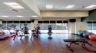 This well-appointed fitness center at 1120 N Division St in Spokane, Washington, offers a comprehensive single-floor workout facility designed to accommodate a wide range of exercise needs. The space features rich hardwood flooring throughout, a suspended tile ceiling with recessed panel lighting that ensures consistent, bright illumination across the entire floor plan.

Large floor-to-ceiling windows line one side of the facility, allowing abundant natural light to fill the space and providing views to the outside, creating an open and energizing atmosphere. The cardio zone is equipped with treadmills, elliptical machines, and stationary bikes, all positioned to take advantage of the window views and wall-mounted televisions for entertainment during workouts.

A dedicated free-weight area includes a full dumbbell rack stocked with a range of weights, along with workout benches and functional training equipment. A mirrored accent wall featuring a bold dark green panel reflects the space and enhances the sense of depth, while stability balls and additional accessories are neatly stored nearby. A pull-up bar and cable training station provide additional strength training options.

The layout is thoughtfully organized, offering distinct zones for cardio, strength, and functional training without feeling crowded. A glass-enclosed room is visible within the space, and posted fitness center rules indicate a professionally managed facility.