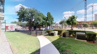 This outdoor dog park, located in Dallas, Texas, serves as a dedicated pet-friendly amenity adjacent to a residential building. The space is enclosed by sturdy black wrought iron fencing with a secure gated entry accented by classic brick pillars, ensuring a safe environment for pets to roam freely. The grounds feature a generous stretch of open green lawn interspersed with patches of natural earth, providing ample room for dogs to run and play. Several mature shade trees are distributed throughout the area, offering welcome relief from the Texas sun and creating a comfortable outdoor setting for both pets and their owners. Green benches are positioned around the perimeter, giving visitors a place to relax while supervising their animals. A curved concrete walkway leads into the space, and waste disposal stations are conveniently placed to encourage cleanliness. The park is well-lit by natural daylight and enjoys views of the surrounding urban Dallas streetscape, with city activity and transit visible beyond the fence line. The adjacent multi-story residential building, clad in brick and siding, provides context for the park's role as a community amenity. Overall, this dog park combines practical design with greenery to create a functional, welcoming outdoor space.