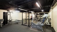 This fitness facility occupies a single floor and is designed to accommodate a range of workout routines. The space features an exposed black-painted ceiling with visible structural beams and ductwork, giving it an industrial aesthetic. Fluorescent strip lighting runs along the ceiling, providing bright, even illumination throughout the room.

The flooring consists of dark rubber matting, which is practical for heavy equipment use and provides traction and impact absorption. The walls are finished in a neutral beige tone, contributing to a utilitarian atmosphere.

The equipment selection covers both strength training and cardiovascular exercise. A prominent power rack stands centrally in the space, paired with a flat bench for free weight exercises. Cardio equipment includes a recumbent stationary bike and at least one treadmill visible toward the rear of the room. Additional multi-use weight machines and other training apparatus are positioned along the back wall. A large mirror panel is mounted on one wall, a standard feature in fitness spaces that aids in form checking during exercise.

Overall, the space presents a functional, no-frills gym environment suited for general fitness use.