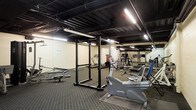 This fitness facility occupies a single floor and is designed to accommodate a range of workout routines. The space features an exposed black-painted ceiling with visible structural beams and ductwork, giving it an industrial aesthetic. Fluorescent strip lighting runs along the ceiling, providing bright, even illumination throughout the room.

The flooring consists of dark rubber matting, which is practical for heavy equipment use and provides traction and impact absorption. The walls are finished in a neutral beige tone, contributing to a utilitarian atmosphere.

The equipment selection covers both strength training and cardiovascular exercise. A prominent power rack stands centrally in the space, paired with a flat bench for free weight exercises. Cardio equipment includes a recumbent stationary bike and at least one treadmill visible toward the rear of the room. Additional multi-use weight machines and other training apparatus are positioned along the back wall. A large mirror panel is mounted on one wall, a standard feature in fitness spaces that aids in form checking during exercise.

Overall, the space presents a functional, no-frills gym environment suited for general fitness use.