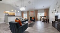 This thoughtfully designed single-floor community space blends contemporary style with welcoming comfort. The main lounge area is anchored by soaring vaulted ceilings and features a striking crystal chandelier that adds a touch of elegance overhead. Light gray hardwood-style flooring flows throughout, tying the open-concept layout together seamlessly. A tile-surround fireplace serves as a focal point, framed by tall windows dressed in warm peach drapes that flood the room with natural light. Plush charcoal velvet sofas and accent chairs adorned with burnt orange pillows create a vibrant, modern seating arrangement, while a sleek coffee table and a high-top bistro set provide flexible gathering options. A decorative wall features sculptural metallic floral art alongside a flat-screen television and media console, adding artistic character to the space. The adjacent kitchen is well-appointed with warm oak cabinetry, granite-style countertops, stainless steel appliances, and a prep island — ideal for events and gatherings. A dedicated beverage station with a coffee maker and microwave offers added convenience. Additional rooms include a flexible multipurpose area with a bulletin board, suitable for meetings or activities. Throughout, chair-rail molding and recessed lighting contribute to a polished, cohesive aesthetic.