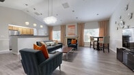 This thoughtfully designed single-floor community space blends contemporary style with welcoming comfort. The main lounge area is anchored by soaring vaulted ceilings and features a striking crystal chandelier that adds a touch of elegance overhead. Light gray hardwood-style flooring flows throughout, tying the open-concept layout together seamlessly. A tile-surround fireplace serves as a focal point, framed by tall windows dressed in warm peach drapes that flood the room with natural light. Plush charcoal velvet sofas and accent chairs adorned with burnt orange pillows create a vibrant, modern seating arrangement, while a sleek coffee table and a high-top bistro set provide flexible gathering options. A decorative wall features sculptural metallic floral art alongside a flat-screen television and media console, adding artistic character to the space. The adjacent kitchen is well-appointed with warm oak cabinetry, granite-style countertops, stainless steel appliances, and a prep island — ideal for events and gatherings. A dedicated beverage station with a coffee maker and microwave offers added convenience. Additional rooms include a flexible multipurpose area with a bulletin board, suitable for meetings or activities. Throughout, chair-rail molding and recessed lighting contribute to a polished, cohesive aesthetic.