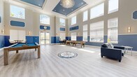 Located at 1-5 Soldier Hill Road in Paramus, NJ, this single-floor clubhouse is a thoughtfully designed community gathering space that combines elegant architectural details with versatile recreational amenities. The main hall commands attention with dramatically soaring double-height ceilings crowned by a coffered tray ceiling finished in a rich blue, from which a striking crystal chandelier is suspended. Light gray hardwood-style flooring runs throughout, and a decorative compass rose mosaic inlay serves as a standout centerpiece feature on the floor.

The two-tone wall treatment — warm beige above and bold blue below — carries consistently throughout the space, creating a cohesive and refined aesthetic. Abundant natural light pours in through tall clerestory windows lining the upper walls, supplemented by elegant wall sconces. French doors provide direct access to the outdoors.

The game area is equipped with a billiards table and shuffleboard table, while a comfortable lounge area features blue leather seating arranged around a linear fireplace set within a marble surround with a wall-mounted television above. A separate kitchenette area includes a bar-height counter with seating, a stainless steel refrigerator, cabinetry, and its own mounted television, making the space well-suited for gatherings and events.