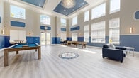 Located at 1-5 Soldier Hill Road in Paramus, NJ, this single-floor clubhouse is a thoughtfully designed community gathering space that combines elegant architectural details with versatile recreational amenities. The main hall commands attention with dramatically soaring double-height ceilings crowned by a coffered tray ceiling finished in a rich blue, from which a striking crystal chandelier is suspended. Light gray hardwood-style flooring runs throughout, and a decorative compass rose mosaic inlay serves as a standout centerpiece feature on the floor.

The two-tone wall treatment — warm beige above and bold blue below — carries consistently throughout the space, creating a cohesive and refined aesthetic. Abundant natural light pours in through tall clerestory windows lining the upper walls, supplemented by elegant wall sconces. French doors provide direct access to the outdoors.

The game area is equipped with a billiards table and shuffleboard table, while a comfortable lounge area features blue leather seating arranged around a linear fireplace set within a marble surround with a wall-mounted television above. A separate kitchenette area includes a bar-height counter with seating, a stainless steel refrigerator, cabinetry, and its own mounted television, making the space well-suited for gatherings and events.