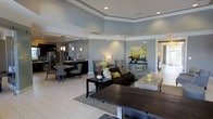 This single-floor space is designed with an open-concept layout that promotes a sense of spaciousness and connectivity between its main living areas. The interior is immediately distinguished by its elevated ceilings, accented with crisp white crown molding and a subtle tray ceiling detail that adds architectural depth to the space.

The living area is anchored by a dark espresso-toned sofa adorned with chartreuse and neutral accent pillows, complemented by a rustic dark wood coffee table and light-toned tile flooring throughout. A soft area rug defines the seating zone, while recessed lighting and a tasteful table lamp contribute to a warm, layered lighting scheme. The walls are finished in a cool, muted blue-gray tone that lends the space a calm and polished ambiance.

Adjacent to the living area, a dining nook and kitchen are visible, featuring dark cabinetry, stainless steel appliances, pendant lighting, and bar-height seating. A console table with decorative accents and a nature-inspired artwork piece add visual interest along the perimeter wall. The overall aesthetic is modern and cohesive, balancing neutral tones with carefully chosen pops of green and yellow throughout.