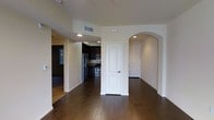 This single-floor space showcases a thoughtful layout with clean lines and a neutral color palette throughout. The main living area is finished with rich, dark hardwood flooring that extends seamlessly into the adjacent spaces, creating a cohesive and polished aesthetic. Recessed lighting illuminates the open layout, highlighting the smooth, light-toned walls that lend the interior a bright and airy feel.

A standout architectural detail is the elegant arched doorway, which adds a classic design touch and provides a graceful transition between the living area and an adjoining hallway. A paneled white interior door nearby offers privacy to an additional room or closet space.

The kitchen is visible from the main living area and is outfitted with dark cabinetry, stainless steel appliances including a refrigerator, stove, and microwave, as well as what appears to be stone or granite countertops. The kitchen flooring features a darker tile in a wood-plank pattern, distinguishing it subtly from the surrounding areas.

The overall ambiance is modern and functional, with practical finishes that balance style and everyday usability. Visible HVAC vents and recessed fixtures speak to a well-maintained, move-in-ready environment.