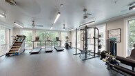 Located at 510 North Wilmington Street in Raleigh, NC, this fitness center offers a comprehensive workout environment thoughtfully spread across a single floor. The main exercise area is bathed in natural light, courtesy of large floor-to-ceiling windows that frame views of surrounding greenery, creating an open and energizing atmosphere. Durable speckled rubber flooring covers the entire space, providing both safety and a clean, professional aesthetic throughout.

The primary workout zone is generously proportioned, accommodating a full row of treadmills, an elliptical trainer, and a stair climber, all positioned to take advantage of the abundant natural light and outdoor views. A heavy-duty power rack with free weights and a bench rounds out the strength training offerings in this area. Ceiling fans and linear LED lighting fixtures ensure comfort and visibility during any workout.

A secondary area features a cable machine and a wall-mounted television, along with relaxed seating — making it suitable for stretching, cooldown, or light exercise. An adjacent room provides space for a stationary cycling bike with an integrated display screen, ideal for focused indoor cycling sessions. A built-in water fountain station with dual dispensers is conveniently located nearby. The overall design is clean, modern, and functional, making this fitness center a well-rounded facility for residents in the Raleigh area.