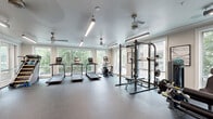 Located at 510 North Wilmington Street in Raleigh, NC, this fitness center offers a comprehensive workout environment thoughtfully spread across a single floor. The main exercise area is bathed in natural light, courtesy of large floor-to-ceiling windows that frame views of surrounding greenery, creating an open and energizing atmosphere. Durable speckled rubber flooring covers the entire space, providing both safety and a clean, professional aesthetic throughout.

The primary workout zone is generously proportioned, accommodating a full row of treadmills, an elliptical trainer, and a stair climber, all positioned to take advantage of the abundant natural light and outdoor views. A heavy-duty power rack with free weights and a bench rounds out the strength training offerings in this area. Ceiling fans and linear LED lighting fixtures ensure comfort and visibility during any workout.

A secondary area features a cable machine and a wall-mounted television, along with relaxed seating — making it suitable for stretching, cooldown, or light exercise. An adjacent room provides space for a stationary cycling bike with an integrated display screen, ideal for focused indoor cycling sessions. A built-in water fountain station with dual dispensers is conveniently located nearby. The overall design is clean, modern, and functional, making this fitness center a well-rounded facility for residents in the Raleigh area.