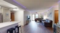 This single-floor residence offers a well-planned layout with a cohesive modern aesthetic highlighted by programmable LED strip lighting that runs along the ceiling perimeter of the main living areas and bedrooms, creating a distinctive, customizable ambiance throughout the space.

The open-concept living room is the heart of the unit, featuring rich wood-style plank flooring, a comfortable seating arrangement centered around a coffee table, and a wall-mounted television. A large floor-to-ceiling window and sliding glass door flood the space with natural light and provide elevated city views. The living area flows seamlessly into a peninsula-style kitchen equipped with light wood cabinetry, a clean white countertop, stainless steel appliances including a refrigerator and range, and track lighting overhead.

The unit includes two bedrooms, each carpeted and accented with LED lighting. The primary bedroom offers ample space for a full-size bed and additional furnishings, while the second bedroom features a wall-mounted TV and a curtained closet. Both bathrooms are finished with warm wood-toned vanity cabinetry, white countertops, large mirrors with overhead lighting, and tile flooring. One bathroom showcases a wide double-sink vanity, adding a practical and stylish touch to the overall design of this contemporary living space.