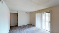 This classic one-bedroom, one-bathroom residence is thoughtfully laid out across a single floor, offering a straightforward and functional living experience. The main living area features soft wall-to-wall carpeting underfoot and a textured ceiling overhead, contributing to a cozy, understated atmosphere. A large sliding glass door fitted with vertical blinds serves as a prominent architectural element, allowing natural light to filter into the space while providing access to the outdoors and views of surrounding greenery. The neutral wall tones create a versatile backdrop well-suited to a variety of furnishing styles. A paneled accent wall adds a subtle touch of classic character to the room. Overhead ventilation and baseboard elements ensure year-round climate comfort. The single-floor design offers ease of access and a seamless flow throughout the unit. Overall, the space presents a clean, move-in-ready environment with classic finishes that provide a solid foundation for comfortable everyday living.