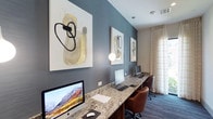 The business center is a thoughtfully designed workspace that balances functionality with contemporary style. The room is anchored by a continuous granite-topped countertop that runs along the length of the space, providing ample room for multiple workstations. Each station is equipped with an iMac computer, keyboard, and mouse, offering a seamless and professional computing experience for users.

The design aesthetic is defined by a striking blue-gray accent wall, which serves as both a visual focal point and a sophisticated backdrop for a curated collection of modern abstract artwork. The paintings, featuring neutral tones of yellow, black, and white, add an artistic touch that elevates the ambiance beyond a typical business center.

Pendant globe lights hang at intervals along the accent wall, casting a warm and focused glow over the workstations. Ergonomic leather chairs in a warm cognac tone provide comfortable seating throughout the space. Underfoot, a patterned blue-gray carpet with subtle linear detailing adds texture and depth to the room.

Natural light filters into the space through a large window dressed with sheer, patterned curtains, offering a soft outdoor view and brightening the overall atmosphere. The combination of modern furnishings, curated art, and natural light creates a productive yet visually refined environment.