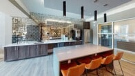 Located in Greenville, SC, this contemporary single-floor community clubhouse is thoughtfully designed to offer a dynamic social hub with a variety of amenity spaces. The open-concept layout is anchored by wide-plank wood-look flooring and a neutral gray color palette that flows seamlessly throughout. Soaring double-height windows in the main lounge area flood the space with natural light and provide views of the outdoor pool area, creating an airy, inviting atmosphere. The lounge is furnished with modern seating and features a shuffleboard table and large-scale artwork, lending a sophisticated yet relaxed feel. A dedicated game area accommodates a full-size pool table, while a charming alcove showcases an oversized wall-mounted Scrabble board with ottomans, adding a playful, interactive touch. The clubhouse kitchen is a standout feature, boasting a striking mirrored subway tile backsplash, dark cabinetry, quartz countertops, stainless steel appliances, and a central island with leather bar seating beneath sleek pendant lighting. A casual dining nook with a round table and curated art wall provides a quieter retreat. Dark textured tile accent walls and recessed lighting add depth and visual interest throughout, completing this well-appointed community gathering space.