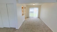 This two-story residential space offers a functional and comfortable layout designed for everyday living. The main living area is open and airy, featuring neutral carpet flooring and light-toned walls that create a clean, welcoming atmosphere. A large sliding glass door floods the space with natural light and provides a direct view of the outdoors, including a green lawn area and neighboring structure, offering a pleasant connection to the exterior environment.

Adjacent to the living area, the kitchen is thoughtfully integrated via a pass-through window, promoting an open feel between the cooking and living spaces. The kitchen is equipped with standard appliances, including a refrigerator and range, and features warm wood-toned cabinetry with light countertops. Overhead shelving provides additional storage options within the kitchen.

Ceiling-mounted lighting fixtures provide consistent illumination throughout the main level. Clean white trim and baseboards frame the rooms neatly, contributing to the overall tidy and well-maintained appearance of the space. With two floors, the layout offers separation between living and private areas, making it a practical and organized home. The overall ambiance is bright, neutral, and move-in ready, suited for those seeking a straightforward and comfortable living environment.